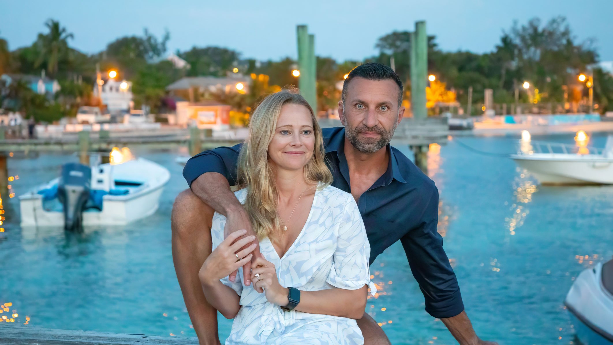 Phil and Erin on a dock at dusk in the Bahamas, harbour lights and boats behind them