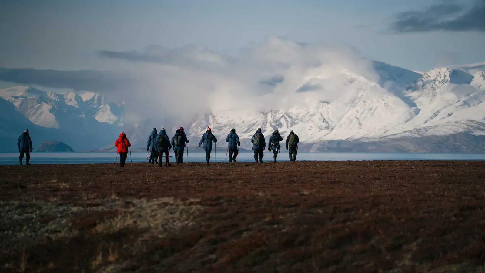 A group of people hike across a grassy coastal field in Greenland
