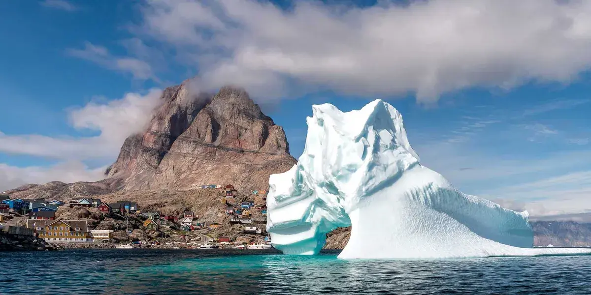 A lone iceberg floats in front of a Greenland mountain village with colorful buildings