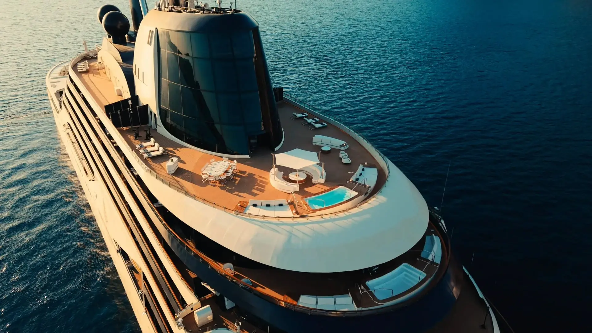 Aerial view of a Four Seasons yacht's top deck at golden hour, with plunge pools, loungers and a glass-domed lounge