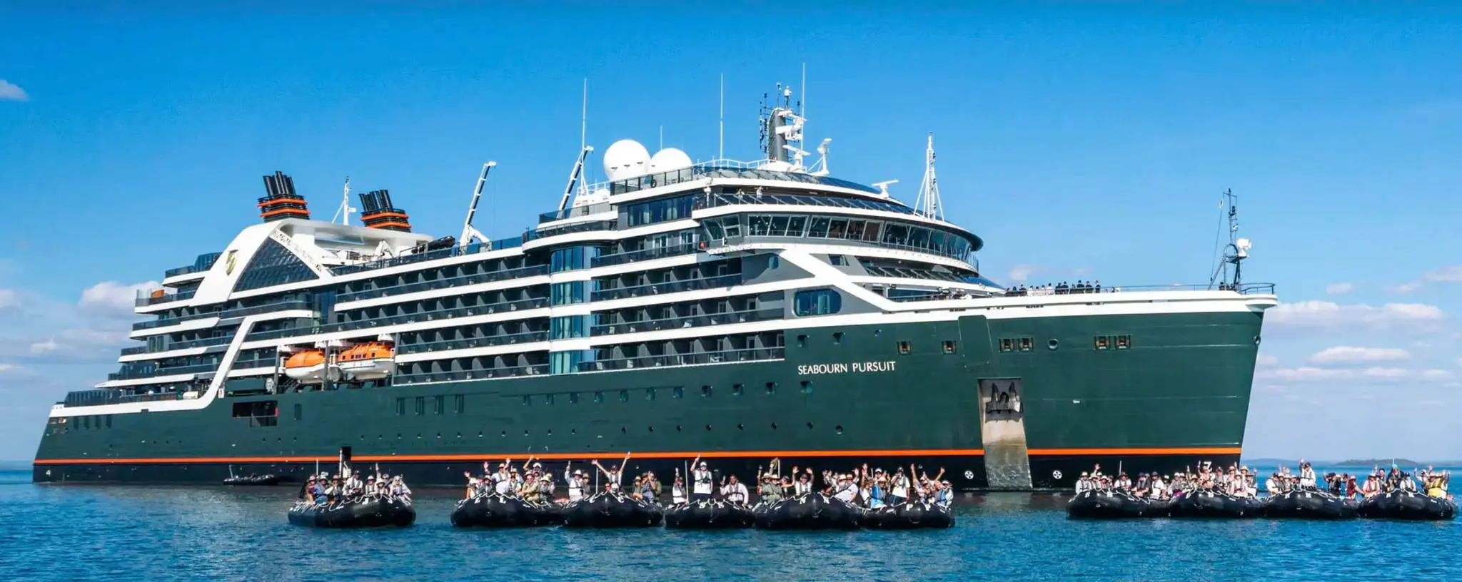 Guests wave from a line of Zodiacs in front of the expedition ship Seabourn Pursuit on calm blue water