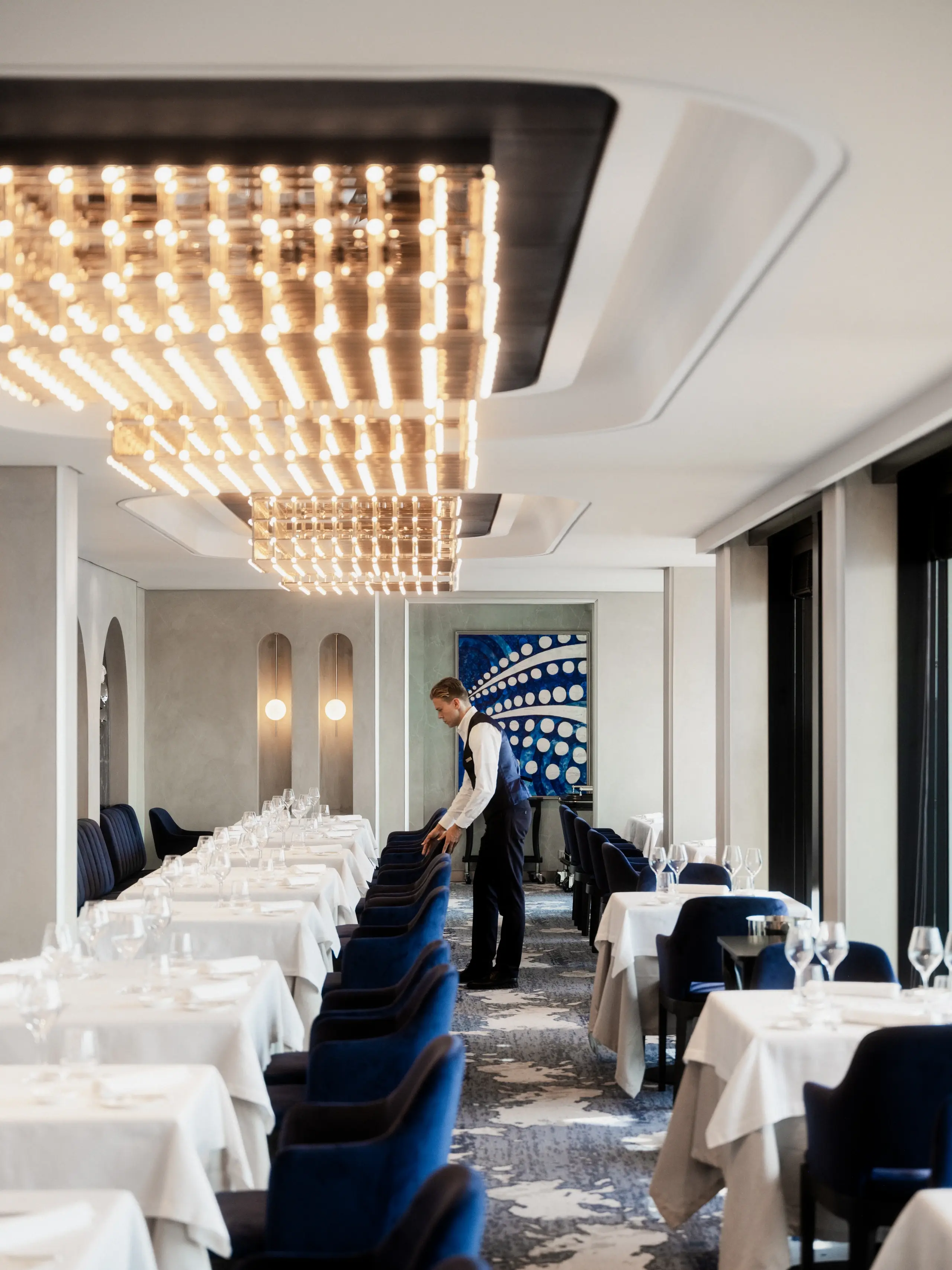 A server sets tables in Anthology, the tasting-menu restaurant aboard Explora III, beneath a glowing light installation
