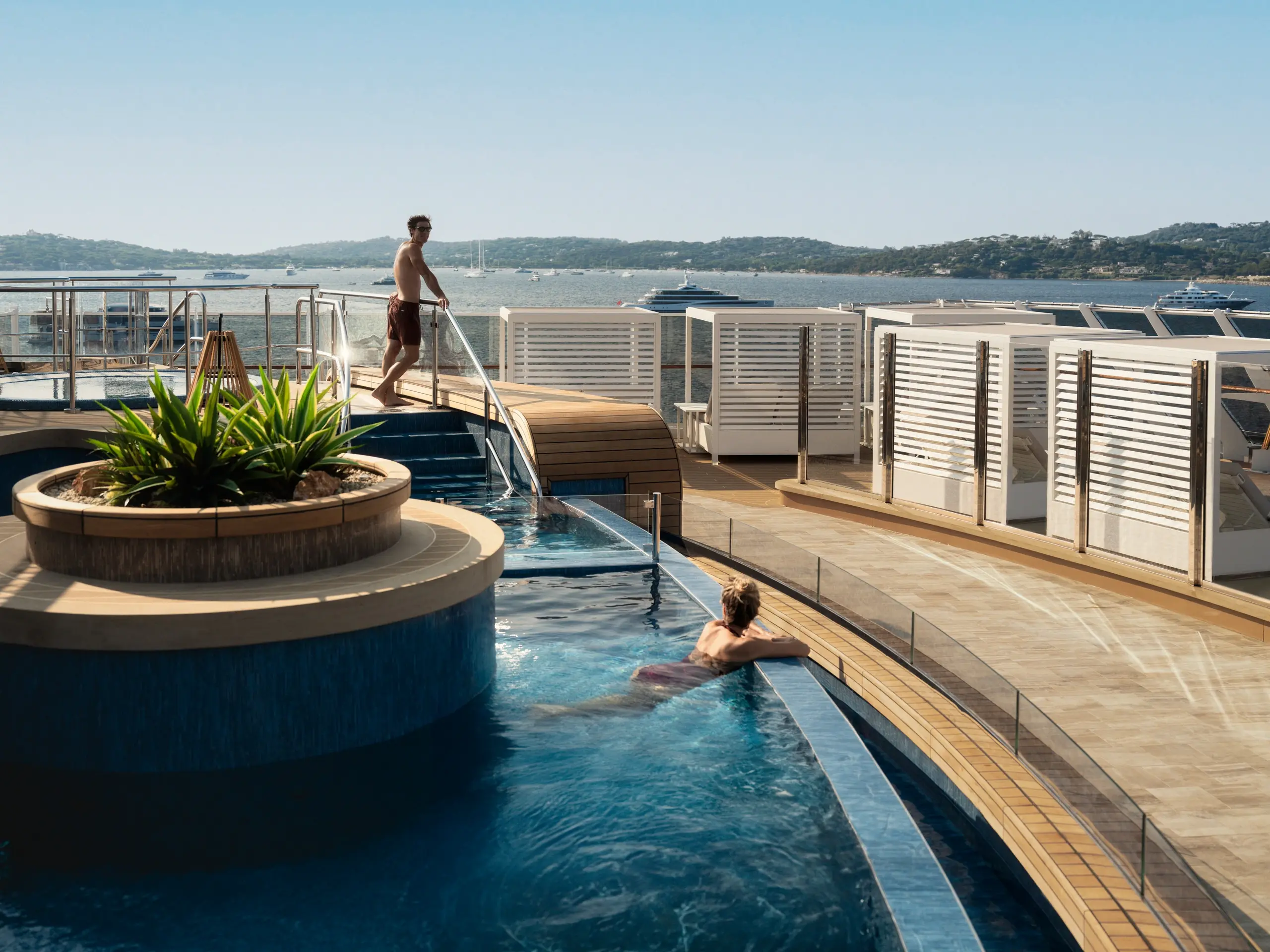A guest leans on the edge of the Helios Pool on Explora III's top deck, with a Mediterranean bay behind