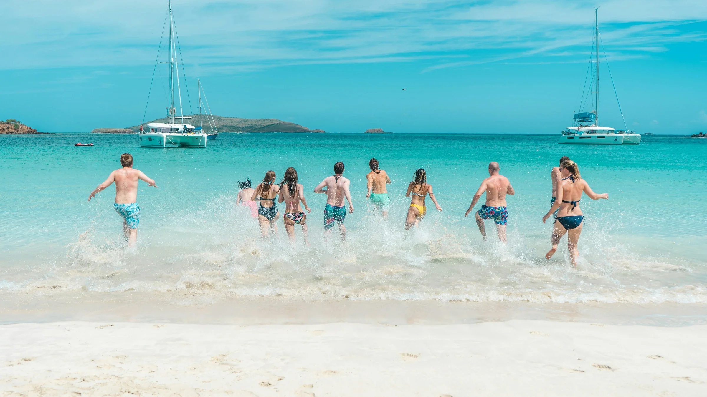 A hosted-journey group runs into a turquoise Caribbean bay, catamarans anchored behind