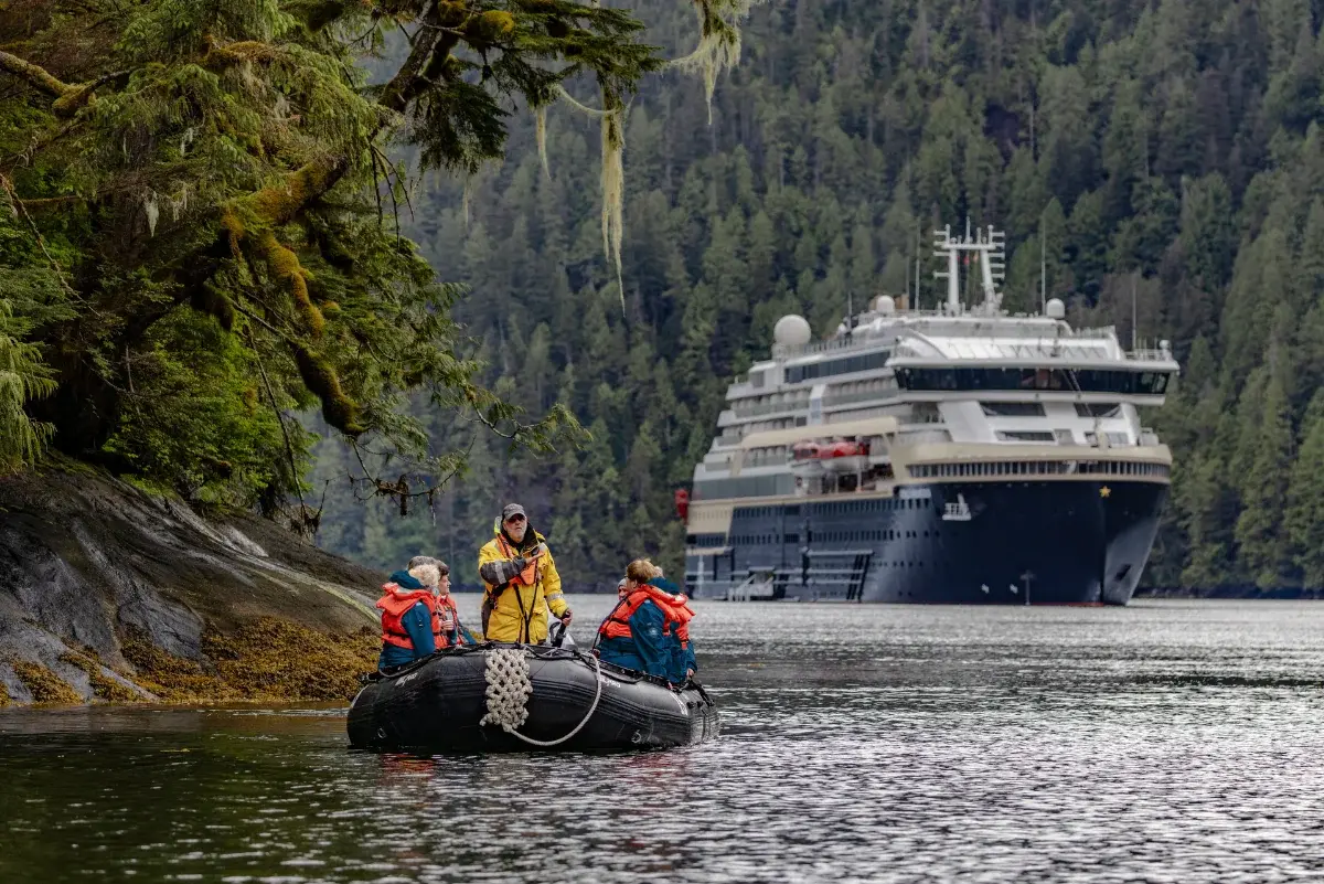 A Zodiac of guests explores a forested Alaskan fjord beside an HX expedition ship